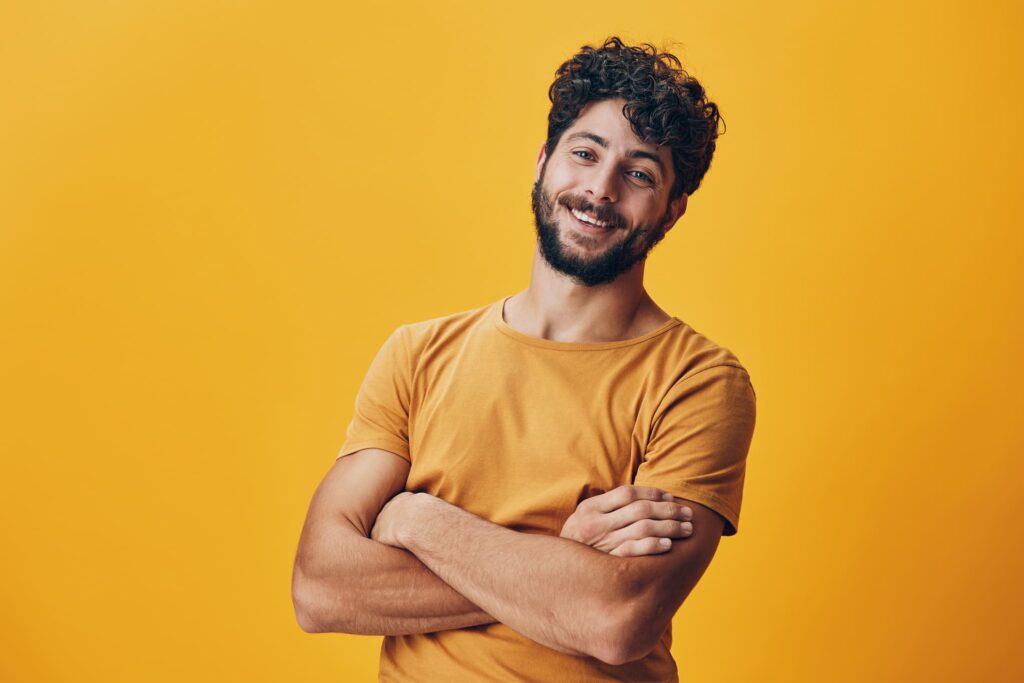 Ein lächelnder Mann mit lockigem Haar und Bart, der ein gelbes T-Shirt trägt, steht vor einem gelben Hintergrund. Er hat die Arme verschränkt und strahlt Zufriedenheit und Freude aus, symbolisch für die Hoffnung und das Glück beim Lotto spielen.