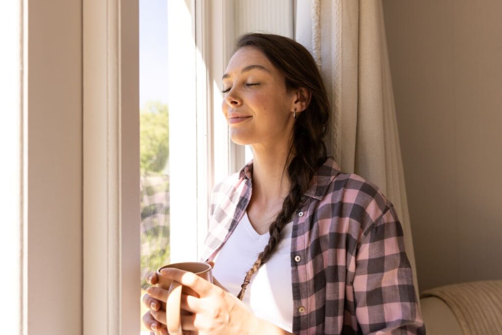 Frau lehnt entspannt am Fenster, trinkt aus einer Tasse und genießt bewusst einen kleinen Genussmoment, passend zur Idee von La Chouffe.